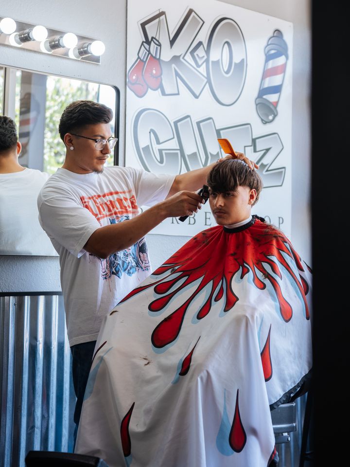 Barber cutting a young client's hair under the K.O. Cutz wall sign